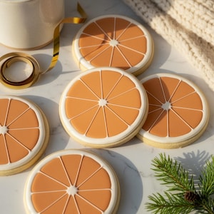 May include: Several orange slice-shaped biscuits with white icing and orange filling are arranged on a white surface. A mug, gold ribbon, and a sprig of pine are also visible. A sweet treat.