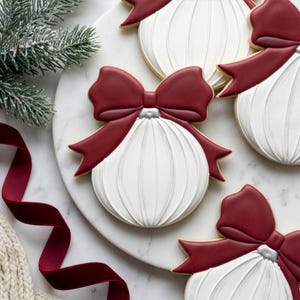 Puede incluir: Primer plano de galletas navideñas con forma de adornos blancos con lazos rojos. Las galletas tienen una superficie texturizada y están decoradas con un acento plateado. Una ramita de hoja perenne y una cinta roja están en el fondo.