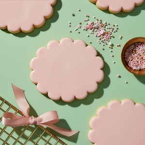 May include: Overhead shot of decorated cookies and sprinkles on a light green surface. The cookies are scalloped-edged and covered in pink icing. A small wooden bowl of sprinkles and a gold-colored cooling rack with a pink ribbon are also present.