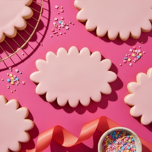 Könnte beinhalten: Draufsicht auf rosa glasierte Kekse mit gewelltem Rand, angeordnet auf einer leuchtend rosa Oberfläche. Bunte Streusel und ein rosa Band sind um die Kekse herum verstreut, zusammen mit einer kleinen Schüssel mit Streuseln und einem goldenen Drahtgestell.