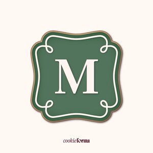 May include: A green cookie cutter featuring the letter "M" in white, set within a decorative frame. The frame has a gold border and white scroll detailing. The brand name "cookieforma" is displayed at the bottom of the image. The overall design is elegant.