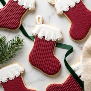 Puede incluir: Galletas con forma de calcetín navideño con glaseado rojo y volantes blancos. Las galletas tienen una textura acanalada y están dispuestas con una cinta verde y una ramita de hoja perenne sobre una superficie de mármol blanco. Dulces festivos.