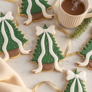 Peut inclure: Biscuits en forme de sapin de Noël avec glaçage vert, des accents blancs et un nœud blanc. Les biscuits ont un motif de panier à la base et sont décorés de points blancs. Une tasse de boisson chaude et un ruban doré sont également présents.