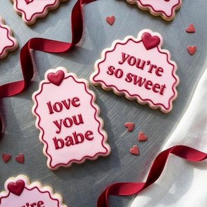 Op de afbeelding: Roze glazuurkoekjes met geschulpte randen en donkerrode tekst met de tekst "love you babe" en "you're so sweet". Kleine hartvormige snoepjes en een rood lint zijn verspreid rond de koekjes op een grijs oppervlak.