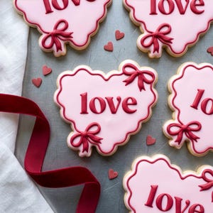 Pode incluir: Biscoitos rosa em forma de coração com a palavra "love" em escrita vermelha. Cada biscoito tem um laço vermelho e uma borda ondulada. Pequenos corações vermelhos espalhados. Uma fita vermelha e um pano branco no fundo.
