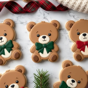 May include: Brown teddy bear-shaped cookies with green, red, and white bow ties. The cookies have a light brown base with darker brown accents and black eyes. A sprig of evergreen and a plaid ribbon are also visible.