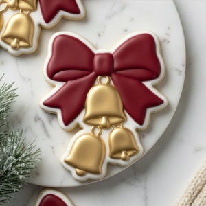 May include: Close-up of a decorated cookie shaped like a bow with three gold bells. The bow is a deep red color with a white outline, and the bells are gold. The cookie is on a white marble surface.