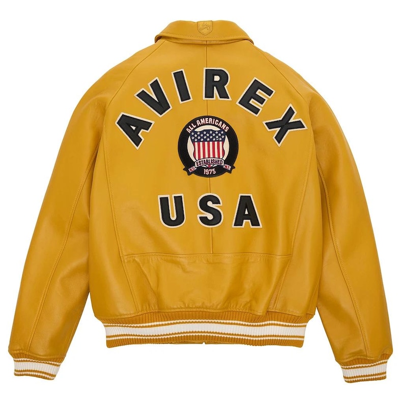 K&ouml;nnte beinhalten: Senfgelbe Lederjacke mit dem Wort "AVIREX" auf dem R&uuml;cken, &uuml;ber einem runden Aufn&auml;her mit den Worten "ALL AMERICANS ESTABLISHED 1975" und einem US-Flaggen-Design. Das Wort "USA" steht darunter. Wei&szlig;-beige gestreifte B&uuml;ndchen.