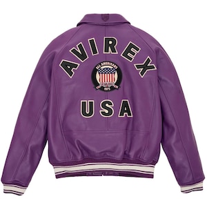 K&ouml;nnte beinhalten: Eine lila Lederjacke mit dem Wort "AVIREX" oben und "USA" unten. Ein kreisf&ouml;rmiger Aufn&auml;her in der Mitte zeigt die amerikanische Flagge und die Worte "ALL AMERICANS ESTABLISHED 1975". Die Jacke hat einen wei&szlig;-schwarz gestreiften Saum.