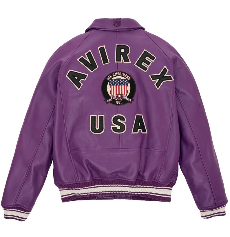 K&ouml;nnte beinhalten: Eine lila Lederjacke mit dem Wort "AVIREX" oben und "USA" unten. Ein kreisf&ouml;rmiger Aufn&auml;her in der Mitte zeigt die amerikanische Flagge und die Worte "ALL AMERICANS ESTABLISHED 1975". Die Jacke hat einen wei&szlig;-schwarz gestreiften Saum.