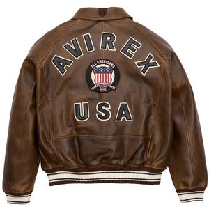 K&ouml;nnte beinhalten: Braune Leder-Bomberjacke mit dem Wort "AVIREX" in wei&szlig;en Buchstaben auf dem R&uuml;cken. Darunter befindet sich ein Aufn&auml;her mit den Worten "ALL AMERICANS ESTABLISHED 1975" und einer Grafik der amerikanischen Flagge. Das Wort "USA" steht darunter.