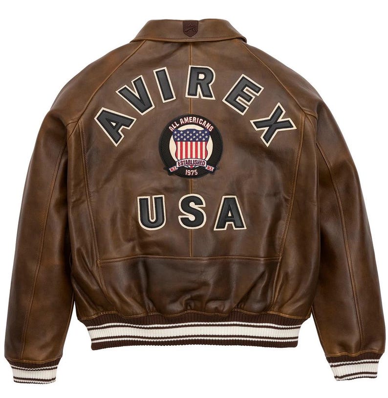 K&ouml;nnte beinhalten: Braune Leder-Bomberjacke mit dem Wort "AVIREX" in wei&szlig;en Buchstaben auf dem R&uuml;cken. Darunter befindet sich ein Aufn&auml;her mit den Worten "ALL AMERICANS ESTABLISHED 1975" und einer Grafik der amerikanischen Flagge. Das Wort "USA" steht darunter.