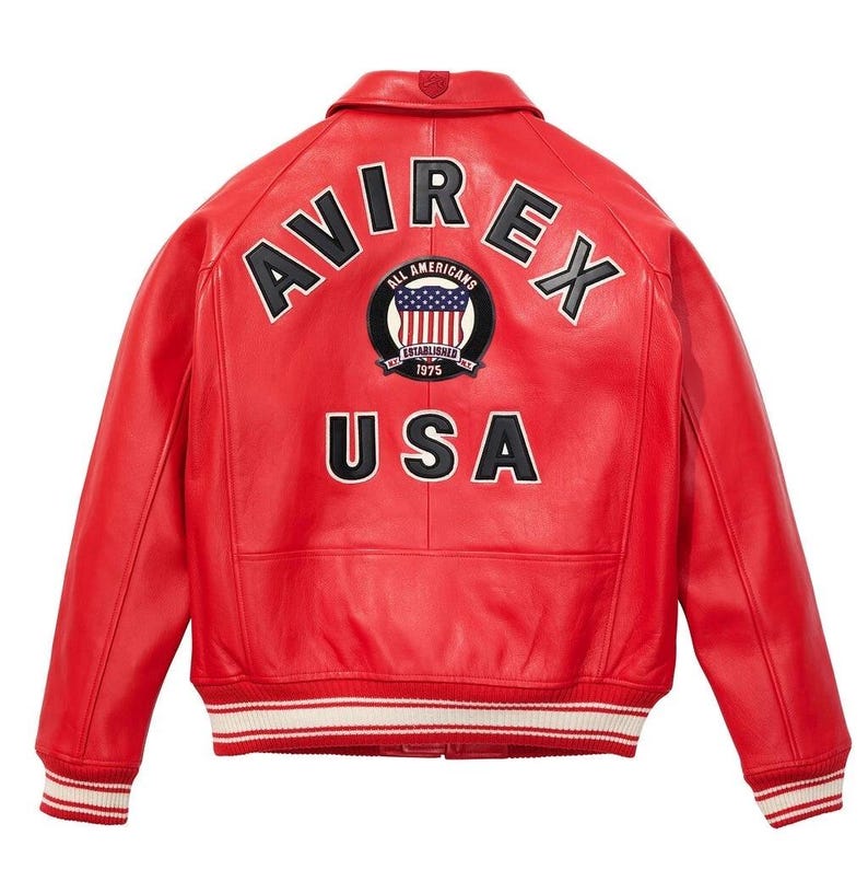 K&ouml;nnte beinhalten: Rote Lederjacke mit dem Wort "AVIREX" in schwarzer Schrift &uuml;ber den Schultern. Darunter steht "USA" ebenfalls in Schwarz. Ein kreisf&ouml;rmiger Aufn&auml;her mit der amerikanischen Flagge befindet sich zwischen den W&ouml;rtern. Die Jacke hat einen wei&szlig;-rot gestreiften Saum.