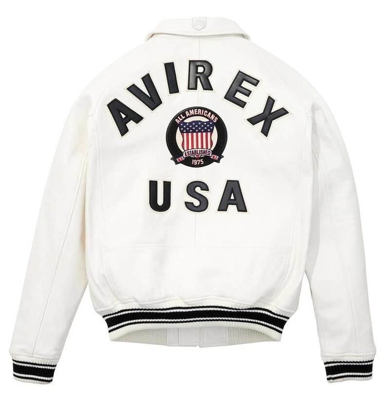 K&ouml;nnte beinhalten: Wei&szlig;e Lederjacke mit schwarz-wei&szlig; gestreiften B&uuml;ndchen und Saum. Auf der R&uuml;ckseite steht "AVIREX" &uuml;ber einem kreisf&ouml;rmigen Emblem mit US-Flaggen-Design und dem Text "ALL AMERICANS ESTABLISHED 1975". Darunter steht das Wort "USA".