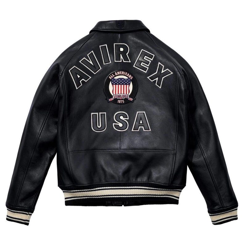 K&ouml;nnte beinhalten: Schwarze Lederjacke mit wei&szlig;er gestickter Schrift "AVIREX" und "USA". Mit einem Aufn&auml;her mit der amerikanischen Flagge und dem Text "ALL AMERICANS ESTABLISHED 1975". Die Jacke hat einen gerippten Kragen, Manschetten und Saum mit cremefarbenen und schwarzen Streifen.