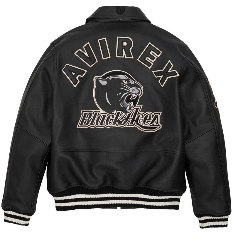 K&ouml;nnte beinhalten: Schwarze Lederjacke mit dem Wort "AVIREX" in wei&szlig;er Schrift &uuml;ber dem oberen R&uuml;cken. Darunter befindet sich eine schwarze Panther-Grafik und die Worte "Black Aces". Die Jacke hat einen gerippten Saum und B&uuml;ndchen mit wei&szlig;en Streifen.