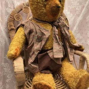 Op de afbeelding: Een vintage teddybeer met gouden vacht, gekleed in een geruit vest, sjaal en bruine shorts. De beer zit in een rieten stoel, wat een nostalgische en charmante scène creëert. De beer heeft een knoop op het vest.