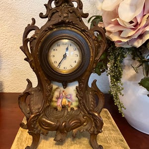 May include: An antique brown metal clock with an ornate design. The clock face is round with black Roman numerals and hands. Below the clock face is a small rectangular painting of two people. The clock is sitting on a light-colored surface.