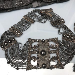 Op de afbeelding: Een donkergrijze kralenketting met meerdere strengen en decoratieve metalen sluitingen. De ketting heeft grote, donkergekleurde edelstenen en ingewikkeld kralenwerk, wat een vintage of antieke uitstraling creëert.