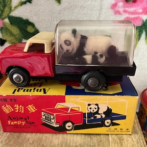 Pode incluir: Um camião de brinquedo vermelho vintage com uma cobertura de plástico transparente, transportando um panda e o seu filhote. O camião está sobre uma caixa colorida com as palavras "Animal Family Van" e uma imagem do camião com um panda. Um brinquedo colecionável.