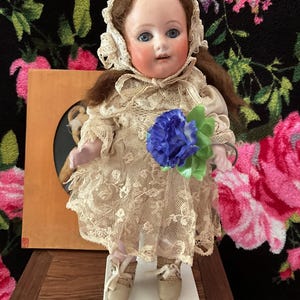May include: A vintage porcelain doll with blue eyes, wearing a lace dress and bonnet. The doll holds a blue flower bouquet. The doll is standing on a white base. The background features a floral pattern.