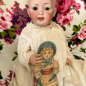 May include: A vintage porcelain doll with a serene expression, wearing a white dress with lace trim. The doll holds a colorful paper cutout of a girl in a dress and hat. The background is a floral pattern with pink and purple roses.