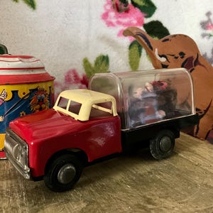 Pode incluir: Um caminhão de brinquedo vintage vermelho e creme com uma parte superior de plástico transparente, rodas pretas e uma cabine creme. Ao lado, um brinquedo de lata colorido com tema de circo e um recorte de elefante marrom.