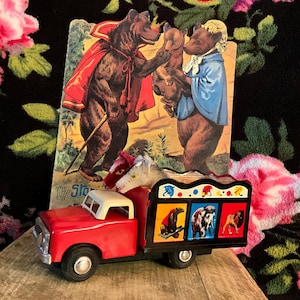 May include: A vintage red toy truck with a white cab and black wheels, carrying two toy horses and a colourful illustrated book. The book depicts bears in a storybook scene. The truck's bed features animal illustrations.