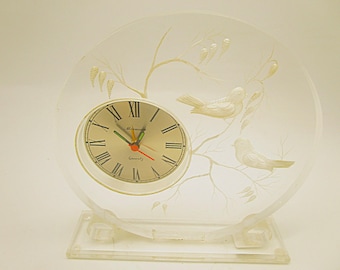 Könnte beinhalten: Eine runde Tischuhr aus klarem Acryl mit einer geätzten Darstellung von zwei Vögeln auf Zweigen. Das Zifferblatt ist silberfarben mit schwarzen römischen Ziffern, grünen Stunden- und Minutenzeigern sowie einem roten Sekundenzeiger. "Quartz" steht darauf.