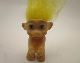 Könnte beinhalten: Eine kleine Troll-Figur aus Kunststoff mit gebräunter Haut, großen spitzen Ohren und leuchtend gelbem, nach oben gestyltem Haar. Die Figur hat eine abgerundete Nase, ein dezentes Lächeln und markante blaue Augen. Sie steht in aufrechter Position.