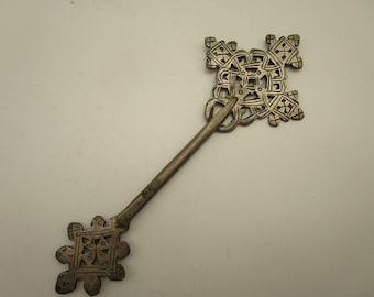 Könnte beinhalten: Eine silberfarbene Metall-Haarnadel mit zwei quadratischen Zierelementen. Jedes Ende zeigt ein komplexes, durchbrochenes geometrisches Kreuzmuster mit eingravierten Linien. Der mittlere Schaft ist ein gerader, glatter Stab.