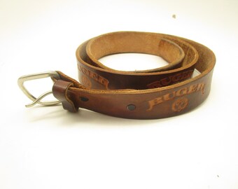 Könnte beinhalten: Ein brauner Ledergürtel mit dem geprägten Schriftzug RUGER und einem kreisförmigen Logo. Der Gürtel verfügt über eine silberfarbene Metallschnalle, eine Lederschlaufe und dunkle Metallnieten, die das Schnallenende des Riemens in aufgerollter Form sichern.
