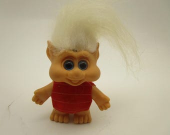 Könnte beinhalten: Eine kleine Troll-Figur mit pfirsichfarbener Haut, großen blauen Augen und einer markanten Nase. Die Figur hat langes, aufrecht stehendes weißes Haar und trägt ein ärmelloses rotes Oberteil mit dünnen horizontalen Streifen sowie vier Zehen.