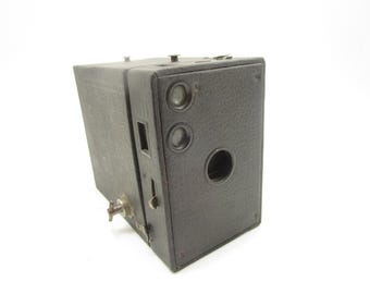 Könnte beinhalten: Eine schwarze Vintage-Boxkamera mit strukturierter Oberfläche, ausgestattet mit einem zentralen runden Objektiv, zwei vertikal übereinander angeordneten kleineren Suchern, einem seitlichen Metall-Drehknopf und einem rechteckigen Auslöser.