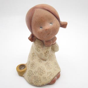 May include: A ceramic figurine of a young girl with brown hair in pigtails, wearing a cream-colored dress with a floral pattern. The figurine has a small, yellow basket at its base. The girl's hands are clasped in front of her.