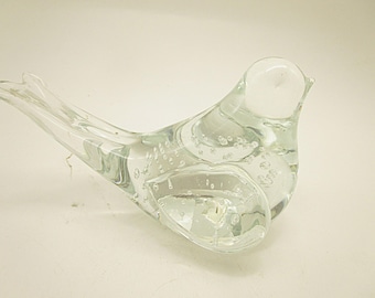Könnte beinhalten: Eine Vogel-Figur aus klarem Glas mit einem glatten, abgerundeten Körper und einem spitzen Schwanz. Das Glas enthält zahlreiche kleine, eingeschlossene Luftblasen in den Flügeln und im Rumpf, die eine texturierte Optik erzeugen.