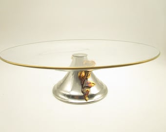 Könnte beinhalten: Ein Tortenständer aus klarem Glas mit einem dünnen Goldrand. Der Ständer verfügt über einen konischen, silberfarbenen Metallsockel, der mit einem skulpturalen, goldfarbenen Rosen- und Blattmotiv verziert ist, das von der Basis bis zur Unterseite reicht.