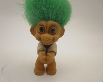 Könnte beinhalten: Eine kleine Trollpuppe mit gebräunter Haut, großen dunklen Augen und einem breiten Lächeln. Die Puppe hat eine hohe, leuchtend grüne Haarsträhne und trägt ein schlichtes, helles Kurzarmshirt. Die Figur steht aufrecht mit nach vorne gestreckten Armen.