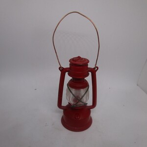 May include: A small, red lantern with a clear glass globe and a copper-colored handle. The lantern has a classic design, with a round base and a chimney-like top. The handle arches over the top, allowing it to be carried or hung.