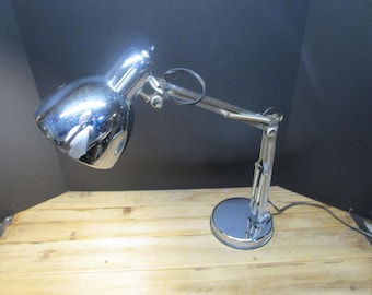 Könnte beinhalten: Eine Schreibtischlampe aus poliertem Chrom mit verstellbarem Gelenkarm, rundem Fuß und kuppelförmigem Lampenschirm. Die Lampe verfügt über ein schwarzes Stromkabel und sichtbare Metallscharniere an den Gelenken der Konstruktion.
