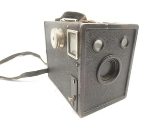 Könnte beinhalten: Eine schwarze Vintage-Boxkamera mit strukturierter Kunstlederoberfläche, ausgestattet mit einem zentralen runden Objektiv, zwei kleinen runden Suchern, einem silbernen Metall-Drehknopf an der Seite und einem schwarzen Trageriemen aus Stoff.