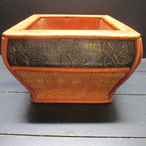 May include: A rectangular wooden planter with a woven base and a dark, decorative band featuring butterfly designs. The planter has a reddish-brown finish and is suitable for indoor or outdoor use.