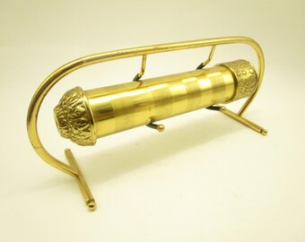 Könnte beinhalten: Ein goldfarbenes Messing-Kaleidoskop auf einem geschwungenen Metallständer. Der zylindrische Körper weist abwechselnd glatte und gebürstete Bänder auf, mit einem geprägten Blumenmuster an einem Ende und einem strukturierten geometrischen Muster am anderen.