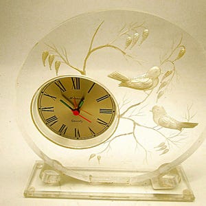 May include: A clear, circular acrylic desk clock featuring an etched design of two birds perched on branches. The clock face is gold-toned with black Roman numerals, black hour and minute hands, and a red second hand. The text "Quartz" is visible.