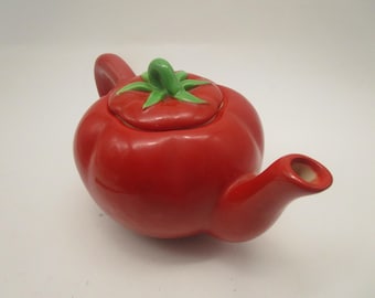 Könnte beinhalten: Eine glänzend rote Keramik-Teekanne in Form einer Tomate. Die Teekanne hat einen abgerundeten Körper, einen geschwungenen Ausguss und einen passenden Deckel, der mit einem grünen, stielartigen Griff versehen ist. Die Oberfläche ist glatt und leuchtend.