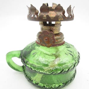 May include: A vintage green glass oil lamp with a handle and a decorative metal top. The lamp has a round base and a textured surface. The metal components show signs of age and wear, adding to its antique charm.
