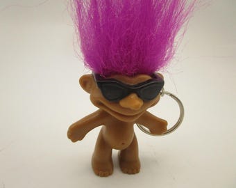 Könnte beinhalten: Eine beigefarbene Troll-Figur als Schlüsselanhänger mit leuchtend violettem Haar und einer schwarzen Kunststoff-Sonnenbrille. Die kleine Figur hat einen breiten, lächelnden Gesichtsausdruck und ist an einem silbernen Metallring befestigt.