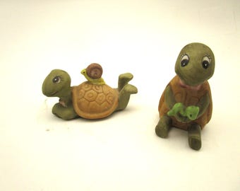 Könnte beinhalten: Zwei Schildkrötenfiguren aus Keramik. Eine Schildkröte liegt auf dem Bauch mit einer kleinen Schnecke auf dem Panzer. Die zweite Schildkröte sitzt aufrecht und hält einen kleinen grünen Gegenstand in den Händen. Beide haben grüne Haut und braune Panzer.
