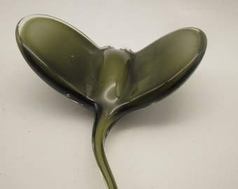 Könnte beinhalten: Eine Skulptur aus durchscheinendem olivgrünem Glas in Form eines Stachelrochens. Das Objekt besteht aus zwei breiten, geschwungenen, flügelartigen Flossen, die von einem zentralen Körper ausgehen, und einem langen, schmalen, spitz zulaufenden Schwanz.