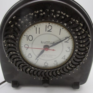 May include: A black electric Remind-O-Timer clock with a white face, black numerals, and a red second hand. The circular dial is surrounded by a black plastic ring featuring numerous small metal pins and numerical markings for time settings.